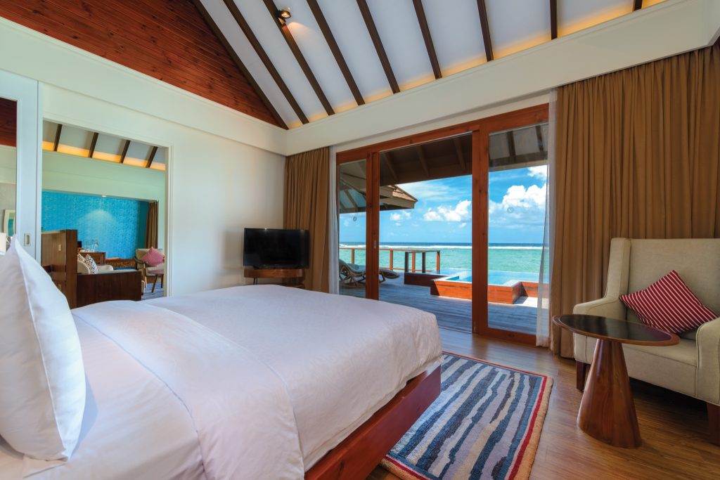 Water Suite Zimmer mit Holzbett, beigem Sessel, rundem Holztisch und Meerblick durch große Fenster