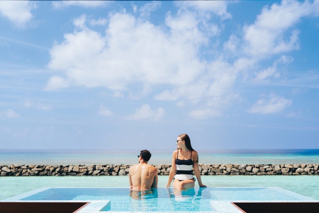Zwei Personen sitzen am Rand eines türkis gefliesten Infinity-Pools mit Blick auf das Meer und eine