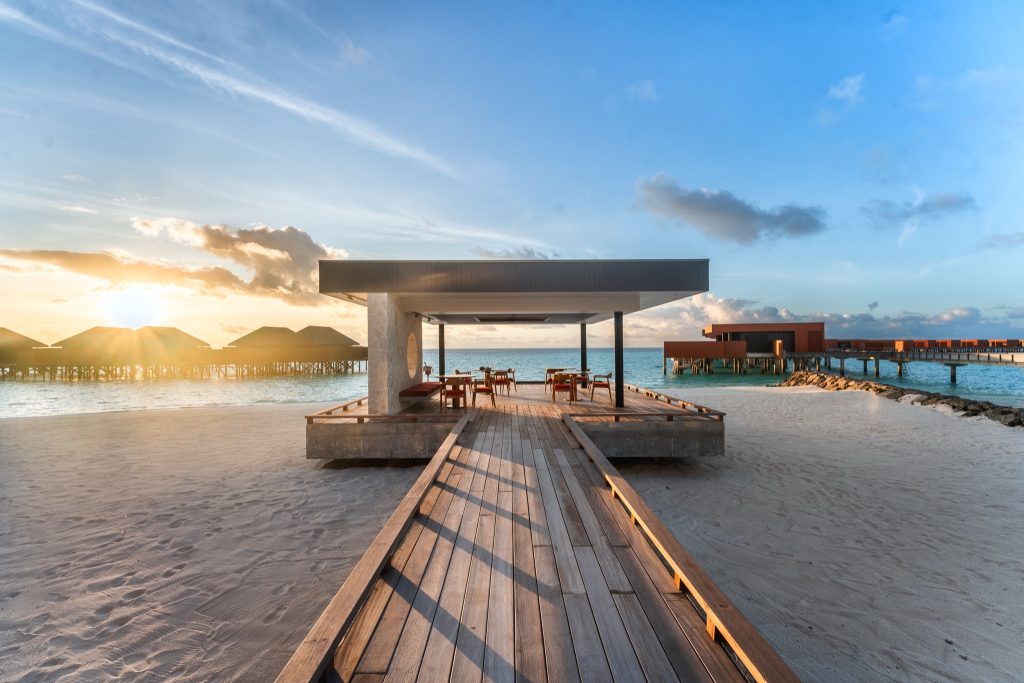 Offener Pavillon mit hellem Holzfußboden und Steinwänden, Wasserbungalows über türkisfarbenem Meer im Sonnenuntergang