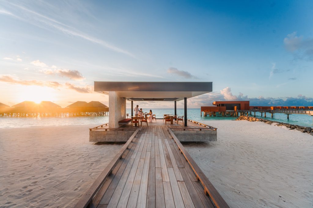 Offenes Pavillon mit Holzmöbeln auf Steg über türkisblauem Wasser am weißen Sandstrand bei Sonnenuntergang