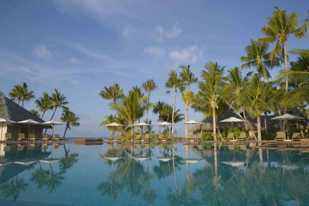 Poolbereich im Veligandu Maldives Resort Island mit Wasser, Holzliegen, Stoffsonnenschirmen und Palmen im Tageslicht