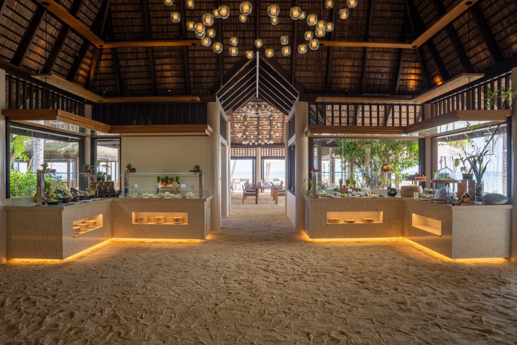 Moderner Buffetbereich im Restaurant Dhonveli mit offenem strohgedecktem Dach, Sandboden und Blick auf tropische Vegetation