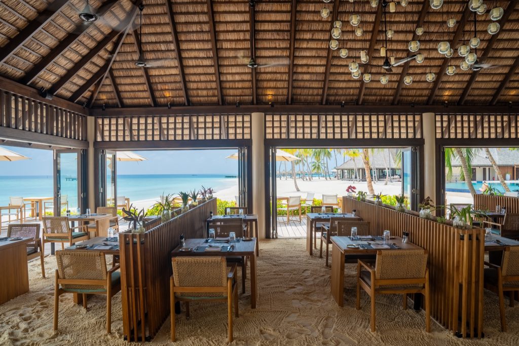 Offenes Restaurant Dhonveli mit Holztischen und Stühlen, Blick auf Strand, Palmen und Terrasse