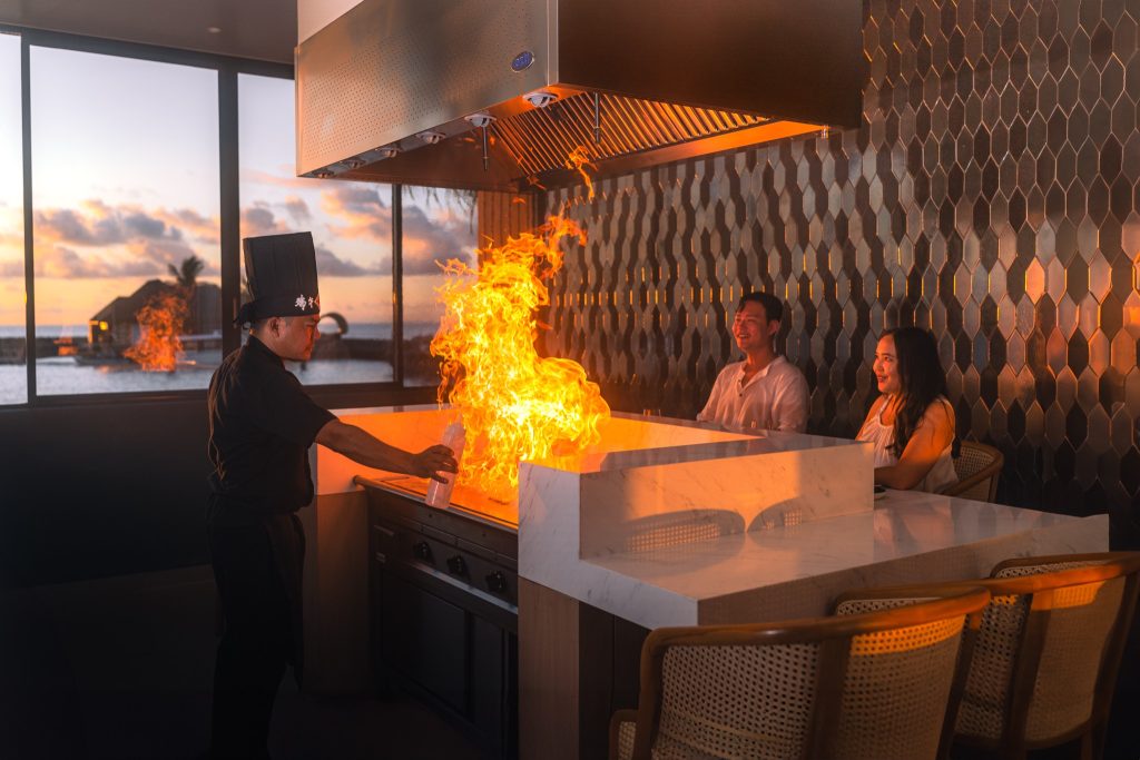 Koch in schwarzer Uniform an Teppanyaki-Herd mit Flamme, zwei Gäste am Marmor-Tresen, dunkle Fliesenwand