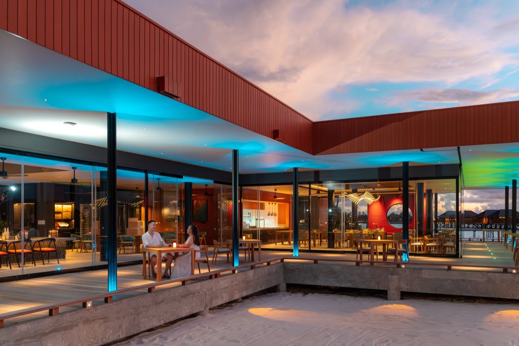Moderne Hotelterrasse des Restaurants Raalhu mit Holztisch, Glaswänden und Sandstrand im Vordergrund