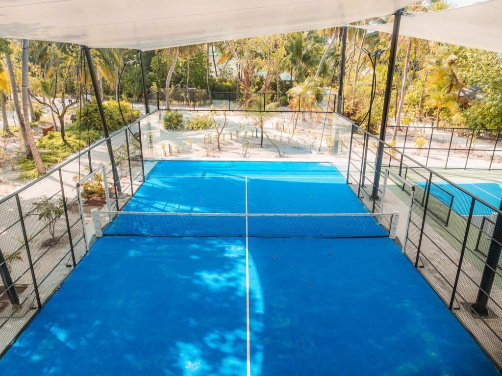 Überdachter Paddletennisplatz mit blauem Verbundboden, Netz, transparenten Wänden und tropischem Garten im Hintergrund