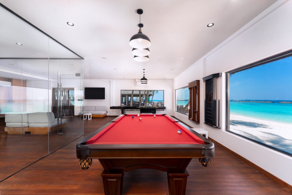 Moderner heller Sport- und Spielraum mit rotem Billardtisch, weißen Wänden, Lounge mit grauen Sofas und Meerblick