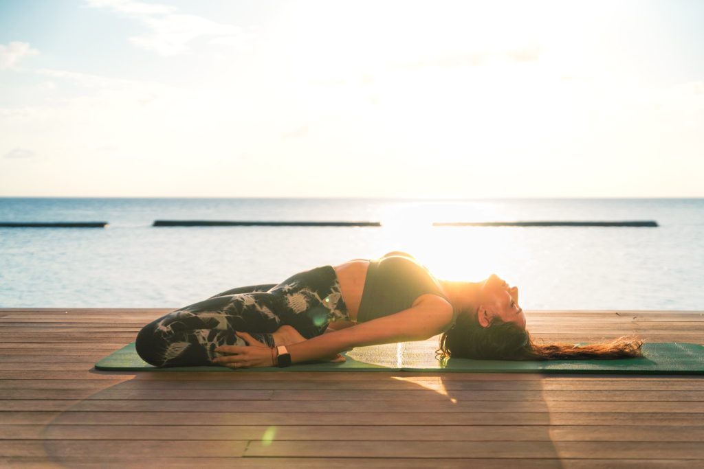 Frau in Rückbeuge auf Yoga-Matte auf Holzdeck mit Meer und hellem Himmel im Hintergrund