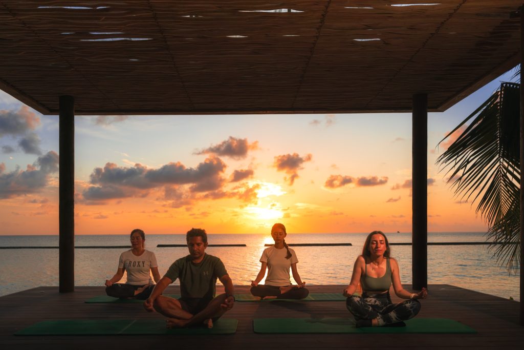 Vier Personen sitzen in Meditationsposition auf einer hölzernen Plattform mit grünem Yogamatten vor Meer und Sonnenuntergang