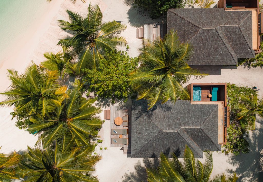 Luftaufnahme von Strandbungalows mit dunklen Schieferdächern, Holzterrassen, Palmen und blauem Pool unter Tageslicht