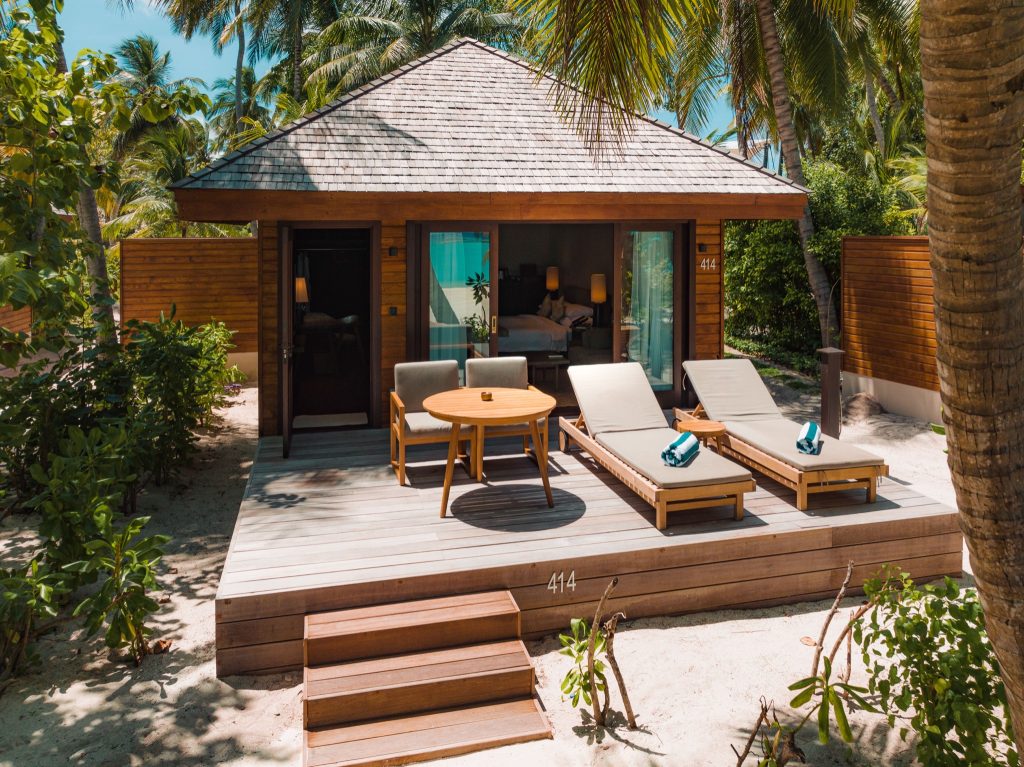 Freistehende Beach Villa mit Holzfassade, Terrasse mit Liegestühlen und Holztisch, umgeben von Palmen