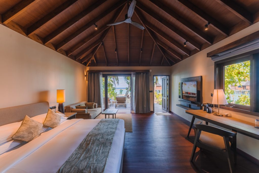 Zimmer der Beach Villa im Veligandu Maldives Resort Island mit Doppelbett, Sofa, Holzbalken und Blick auf Balkon mit Palmen.