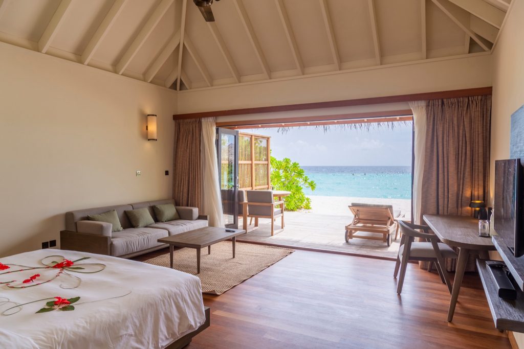 Geräumiges Zimmer der Jacuzzi Beach Villa mit Holzfußboden, großen Fenstern und Blick auf Terrasse und Meer