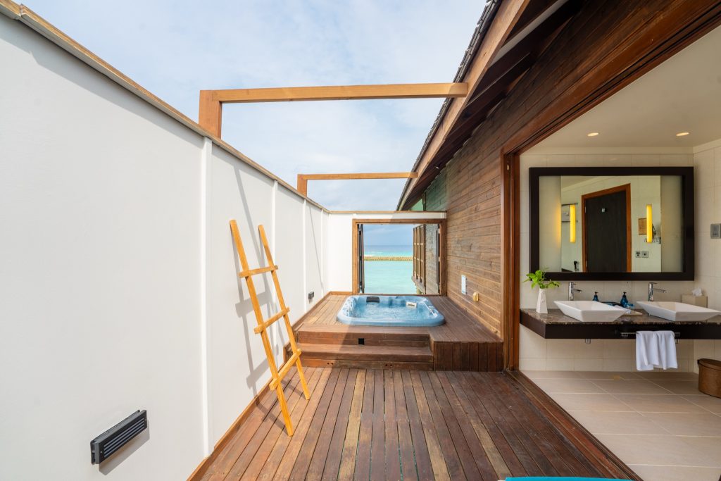 Terrasse der Ocean Jacuzzi Villa mit Holzdeck, Whirlpool, offenen Waschbecken und Einblick ins Badezimmer
