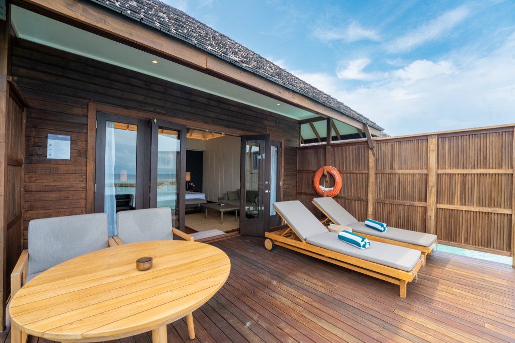 Hotelterrasse mit dunklem Holz, runder Holztisch, zwei gepolsterte Stühle und Liegestühle mit grauen Auflagen