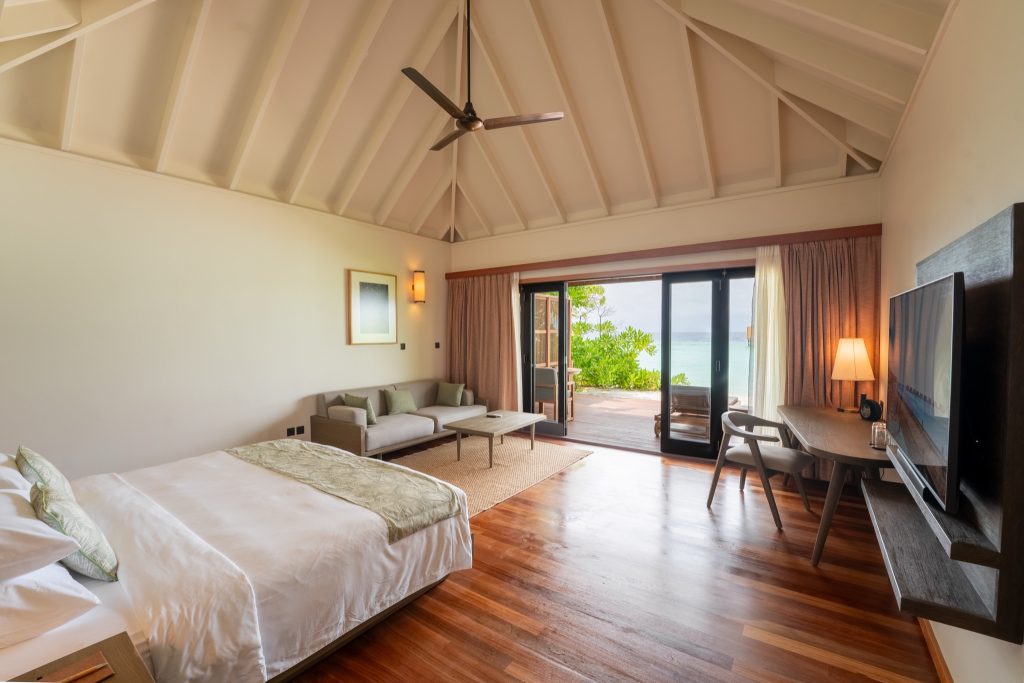 Hotelzimmer der Sunset Jacuzzi Beach Villa mit großem Bett, Sitzecke, Holzboden und Meerblick durch Glastür