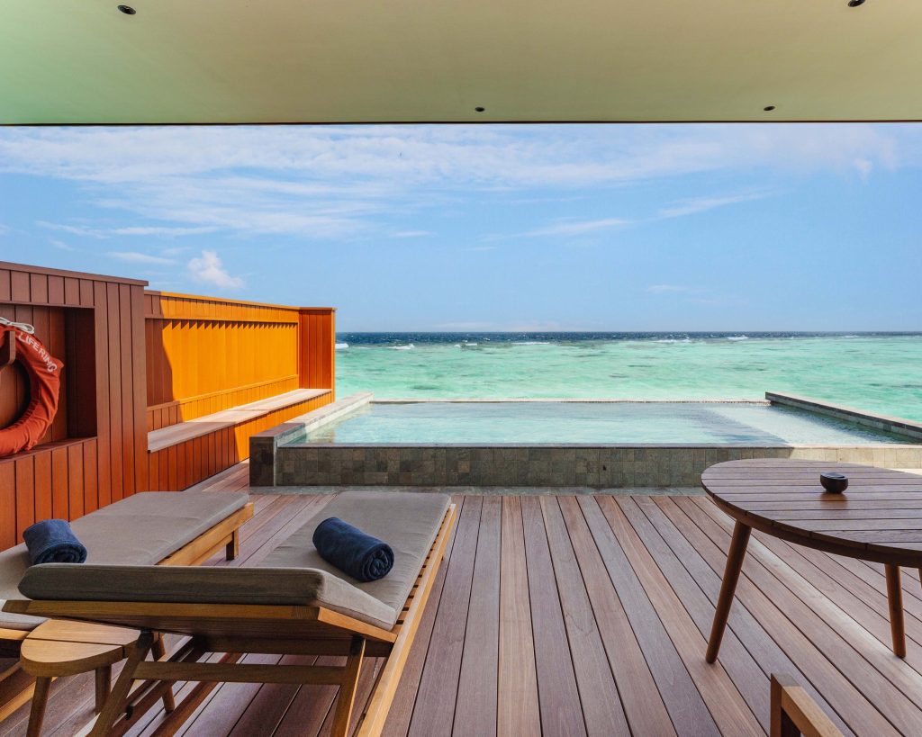 Terrasse eines Hotelzimmers mit Holzpanelen, zwei Liegestühlen, rundem Holztisch und kleinem Tauchbecken mit Meerblick