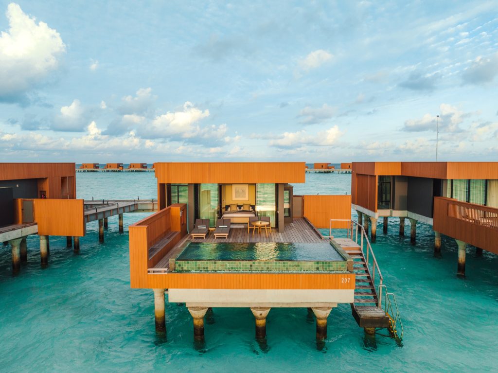 Moderne Wasservilla auf Stelzen mit Holzfassade, Infinity-Pool und Liegestühlen über türkisfarbenem Wasser