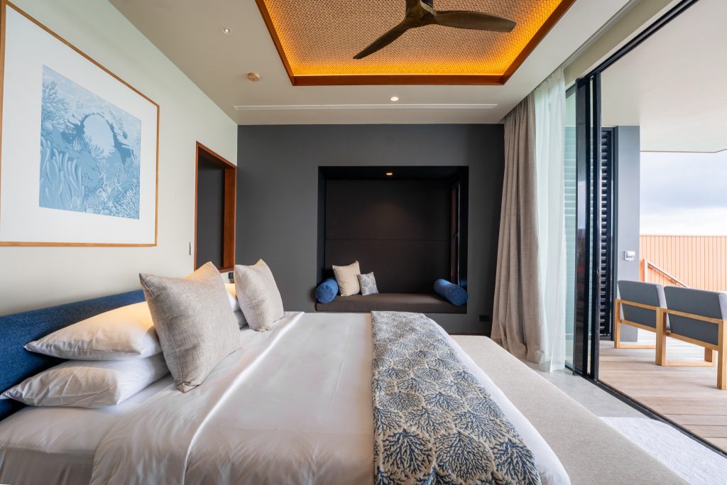 Modernes Hotelzimmer mit Doppelbett, blauem Kunstwerk, großem Fenster zum Balkon mit Holzmöbeln und Decke mit Ventilator