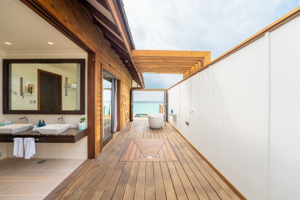 Offener Gang in Zimmer mit Badezimmer links, zwei Waschbecken, freistehender Badewanne und Blick auf das blaue Meer