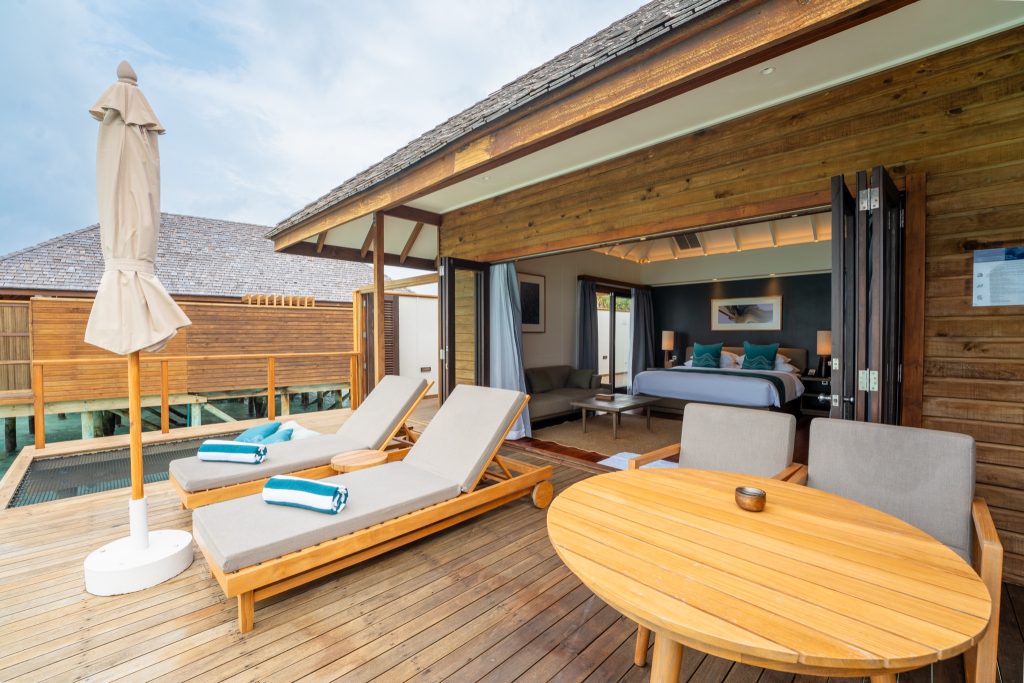 Terrasse eines Hotelzimmers mit zwei gepolsterten Liegen, Holztisch, Sesseln und offenem Wohnbereich mit Bett und blauen.