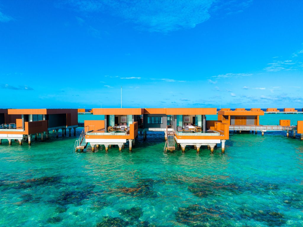 Über Wasser gebaute Two Bedroom Ocean Pool Villas mit oranger Fassade auf Stelzen im türkisfarbenen Wasser bei klarem Himmel