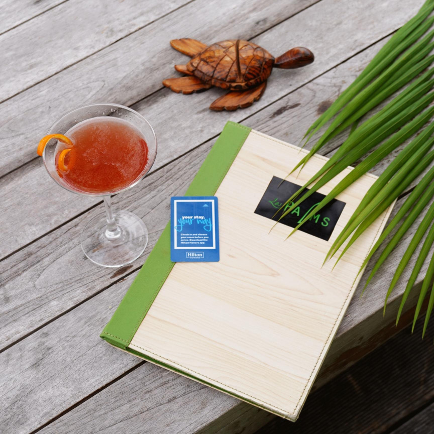 Cocktail im Martini-Glas auf hellem Holztisch mit hölzerner Schildkröte, grüner Mappe und blauer Hilton-Karte unter Palmen