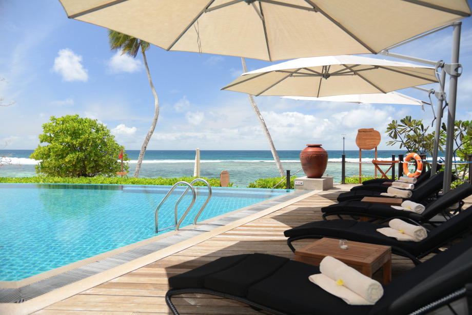Außenpool mit blauen Wasser, Holzterrasse, schwarzen Sonnenliegen, beigen Sonnenschirmen und Meerblick auf den
