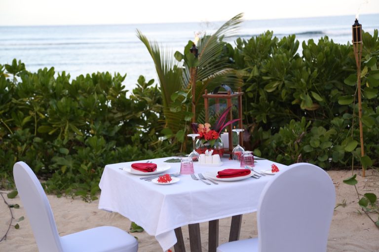 Tisch mit weißer Tischdecke, zwei mit weißem Stoff bezogene Stühle, rote und rosa Blumendekoration am Strand