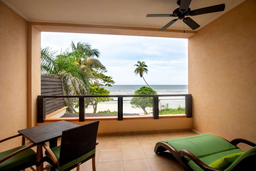 Balkon des Deluxe Ocean View Zimmers mit Holztisch, Stühlen, Liege und Meerblick eingerahmt von Palmen