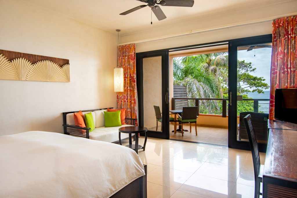 Zimmer deluxe-ocean-view mit großem Bett, Holztisch, Sofa mit bunten Kissen und Balkon mit Palmenblick
