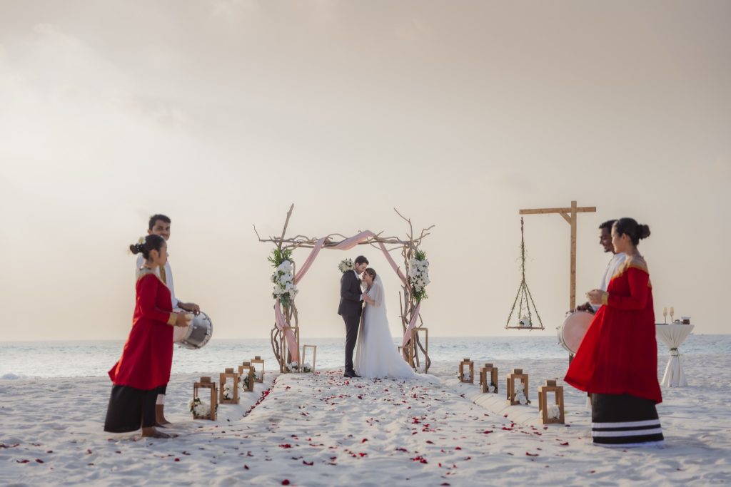 Paar bei Strandhochzeit unter Holzbogen mit Blumen, weißem Teppich und Laternen am Sandstrand mit Meer im Hintergrund
