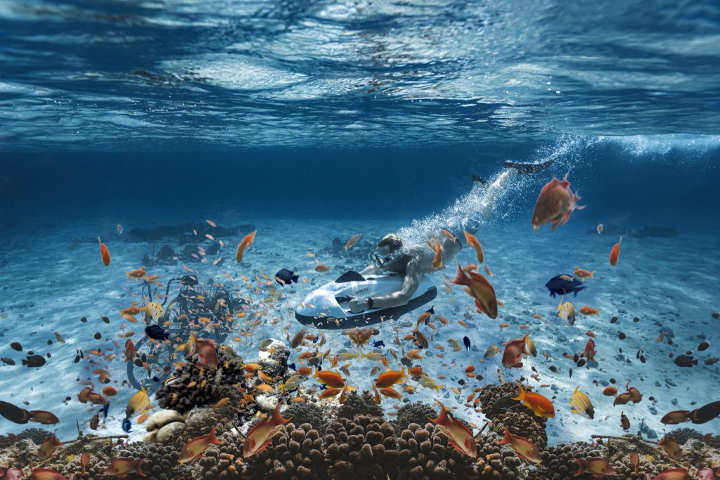 Person mit Unterwasser-Scooter schwimmt über sandigem Meeresboden zwischen Korallen und bunten Fischen