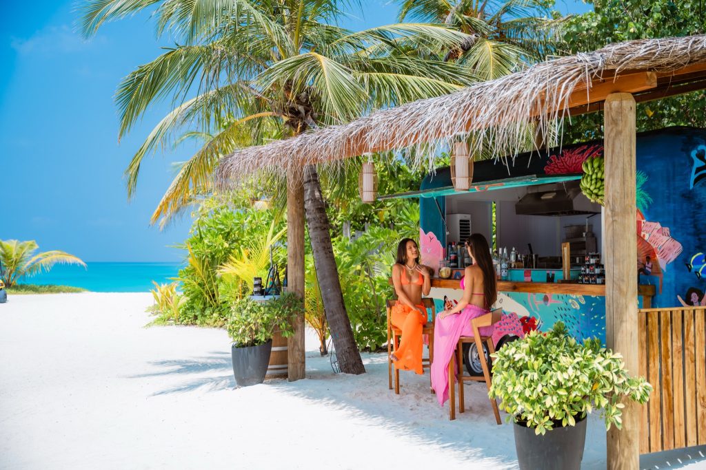 Strandbar aus Holz mit bunten Mustern, umgeben von Palmen und weißem Sand am Strand