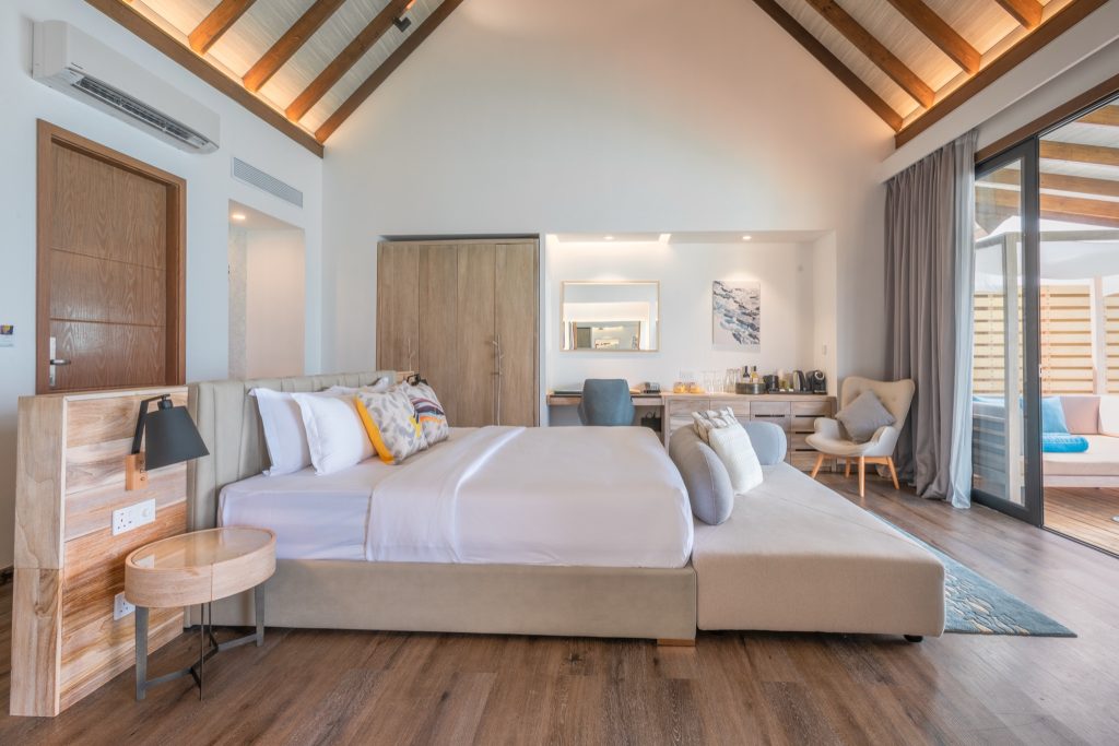 Hotelzimmer mit großem Bett, Holzkopfteil, rundem Holztisch, schwarzer Lampe und Glasschiebetür zum Balkon