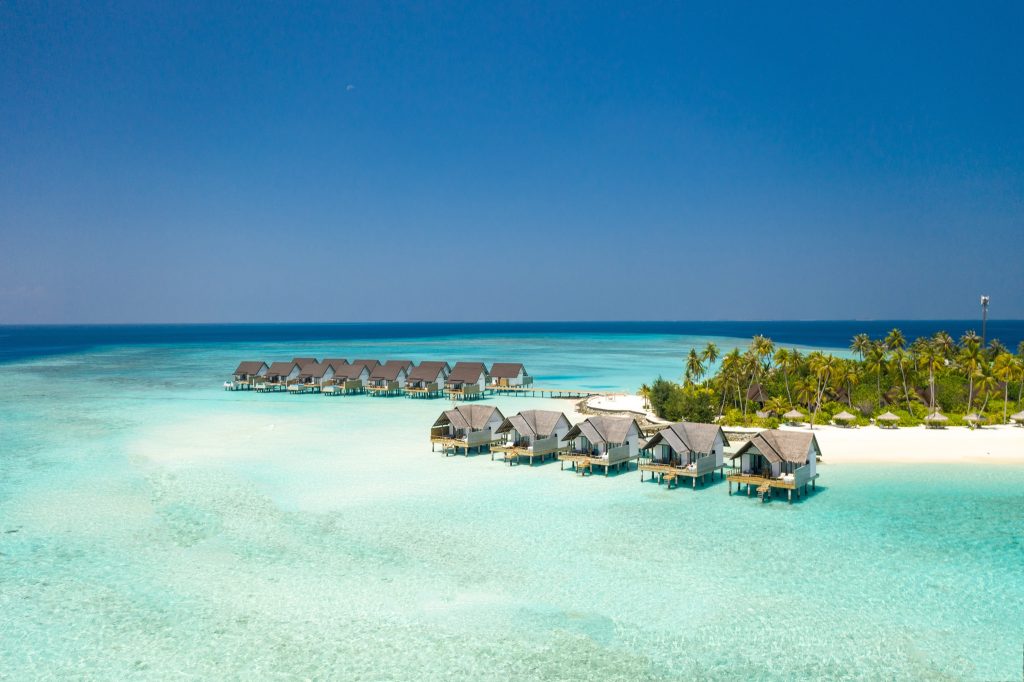 Überwasser-Bungalows auf Stelzen über türkisblauem Wasser mit weißem Sandstrand und Palmen im Hintergrund auf den