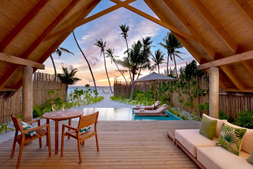 Offene Terrasse mit Holzdielen, Holztisch, Sofa, kleinem Pool und Sandstrand mit Palmen im Hintergrund bei Sonnenuntergang