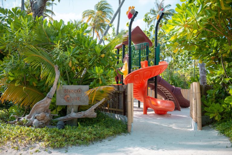 Kinderspielplatz Kokko Club mit orangefarbener Rutsche, Sandboden und tropischer Vegetation auf den