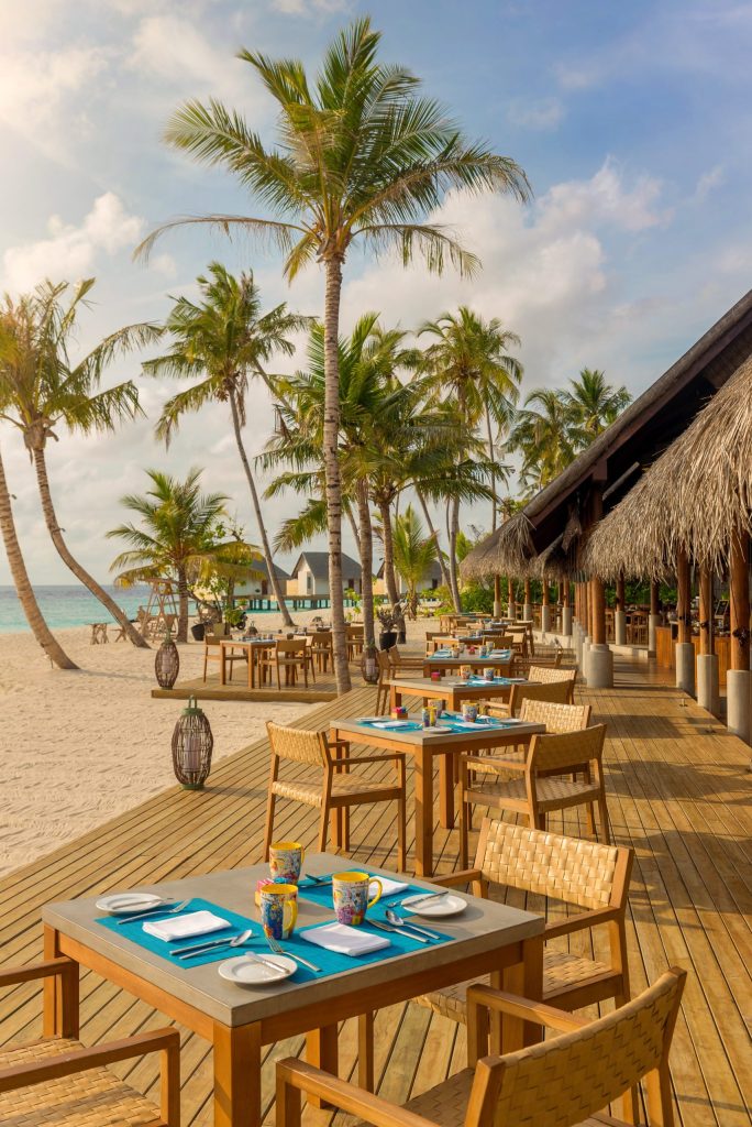 Gedeckte Tische mit hellen Holzstühlen auf Holzterrasse am Strand, Palmen und Meer im Hintergrund