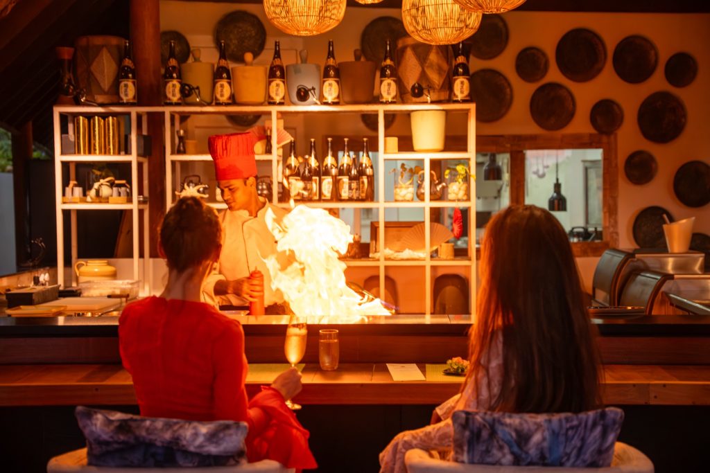 Koch am Teppanyaki-Tisch mit Flamme, zwei Gäste auf gepolsterten Stühlen, warme Beleuchtung und dekorierte Wände