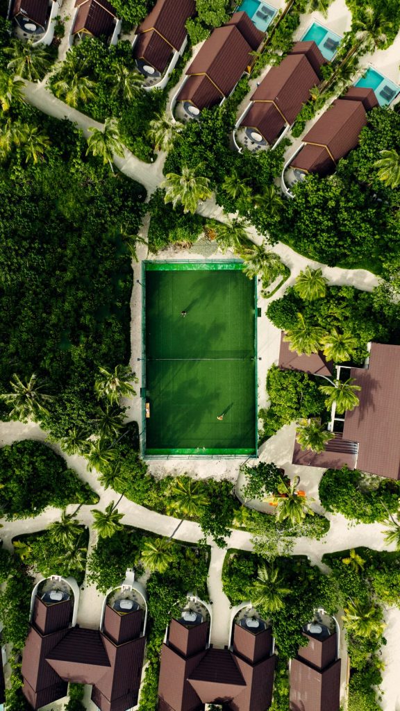 Luftaufnahme eines Tennisplatzes mit zwei Spielern, umgeben von grünen Bungalows und Palmen im Fushifaru Maldives Resort