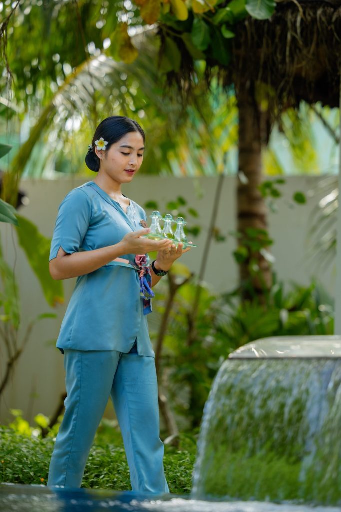Person in blauem Outfit trägt Glasplatte mit Glaskaraffen vor tropischer Vegetation im Heylhi Spa Fushifaru Maldives