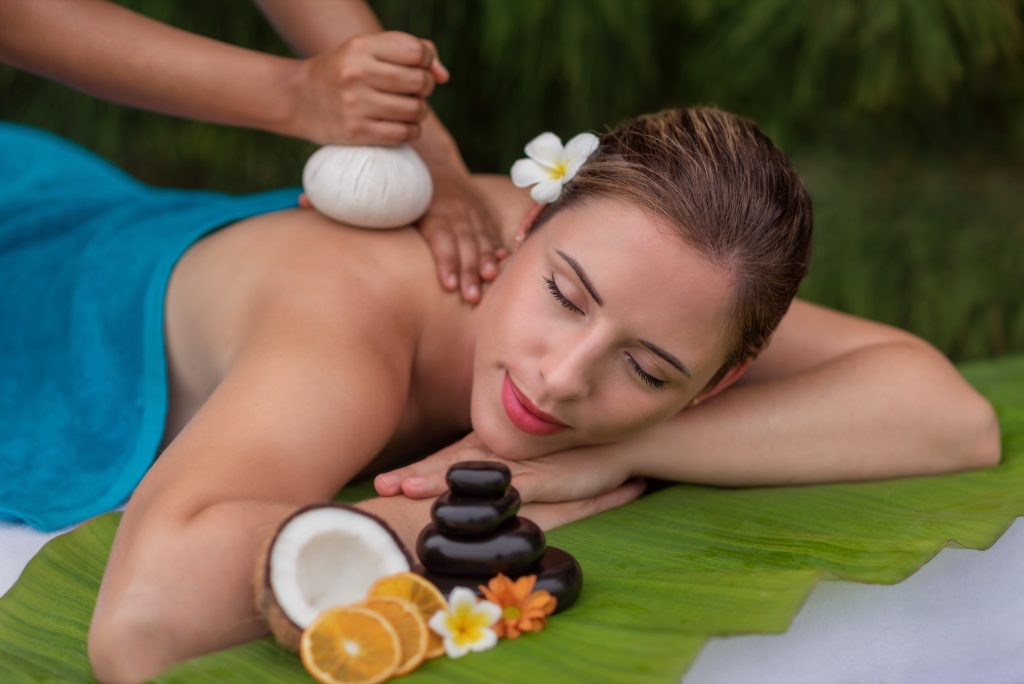 Frau erhält Rückenmassage mit weißem Kräuterbeutel auf grüner Blättermatte im Heylhi Spa Fushifaru Maldives