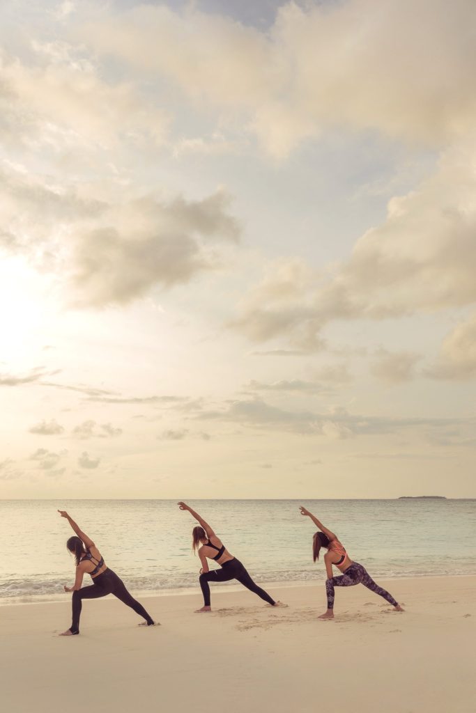 Drei Personen beim Yoga am hellen Sandstrand mit ruhigem blauem Ozean und pastellfarbenem Himmel im Hintergrund