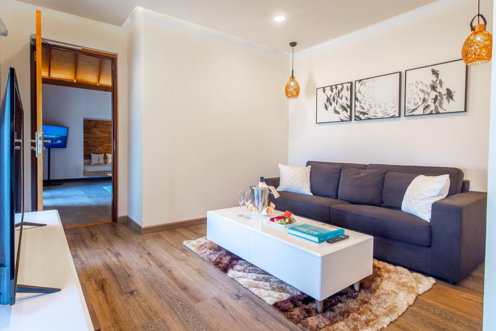 Wohnraum der Beach Duplex Villa im Fushifaru Maldives mit grauem Sofa, weißem Couchtisch, Holzboden und goldenen Hängelampen