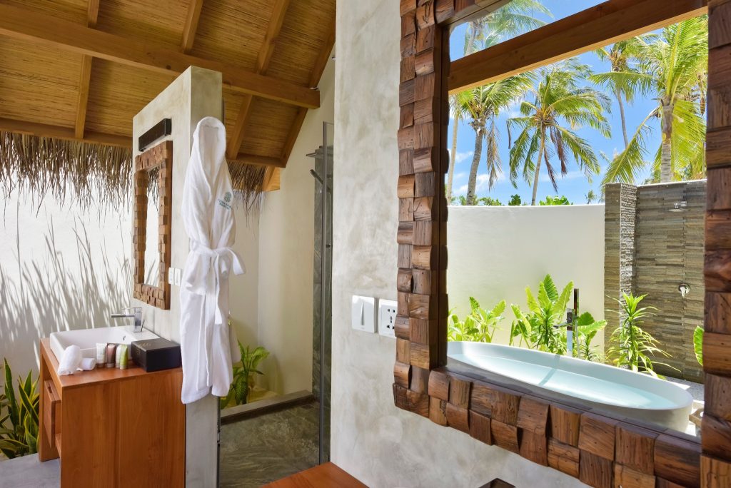 Badezimmer der Beach Villa im Fushifaru Maldives mit freistehender Badewanne, Holzdecke, Steinwänden und Blick auf Palmen