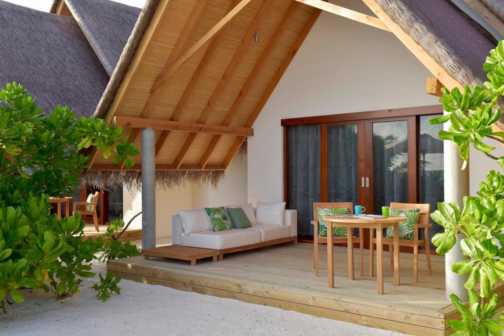 Terrasse im Fushifaru Maldives mit Strohdach, Sofa mit grünen und weißen Kissen, Holztisch und Stühlen aus hellem Holz