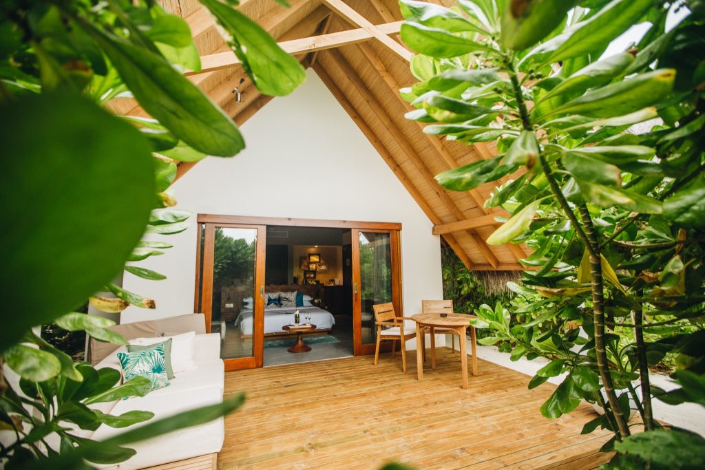 Terrasse der Beach Villa im Fushifaru Maldives mit Holztisch, weißem Sofa und grünen Kissen vor offenem Zimmer