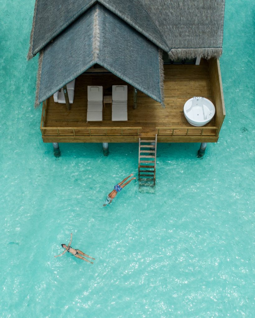 Wasserbungalow auf Holzpfeilern über türkisfarbenem Meer mit Holzterrasse, Whirlpool und drei schwimmenden Personen