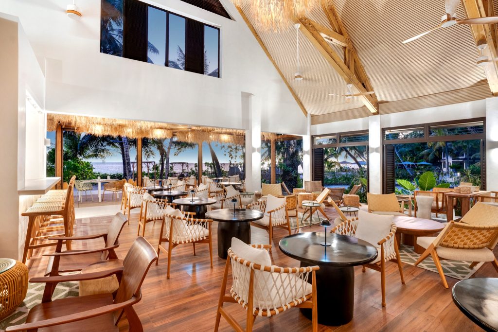 Helle Bar Lounge der Indian Ocean Lodge mit Holz- und Rattanmöbeln, großen Glasfenstern und Blick auf Strand und Grün
